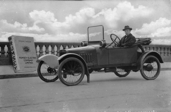 View of an man sitting in an electric car by Rumsey, founded in 1895. A Rumsey Electric Company Limited General Catalog is depicted alongside the vehicle.