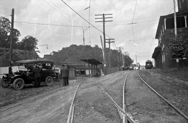 View of trolley cars and a depot. Three men are standing on the left near parked cars. In the background is a roundhouse.