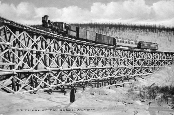 View of a railroad bridge with a train crossing it. A man can be seen standing on the ground next to the bridge. Caption reads: "R.R. Bridge At Fox Gulch, Alaska."