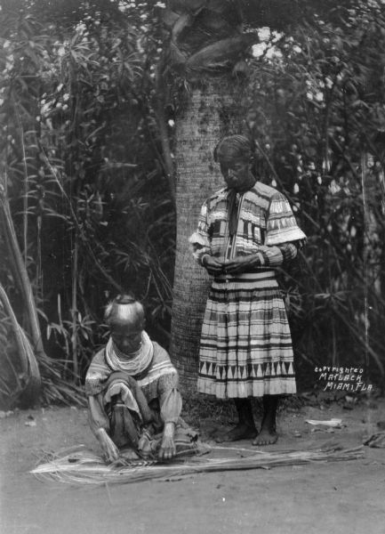 A view of two Seminole Native Americans, one person standing and another person crouching near the ground working on something unidentified.