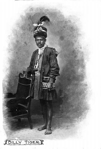 Full-length portrait of Billy Tiger, a Seminole Native American, posed in full regalia standing next to a chair.