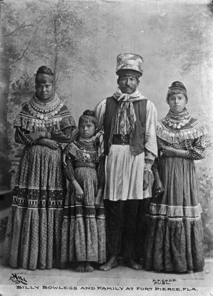 Group portrait in front of a painted backdrop of Billy Bowlegs and his family. They are all wearing native Seminole clothing. Caption reads: "Billy Bowlegs and Family at Fort Pierce, FLA."