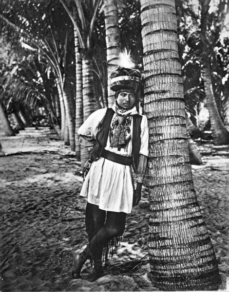A Seminole Native American man wearing native Seminole clothing, leaning against a palm tree, most likely in Palm Beach, Florida.