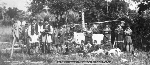 A large Seminole Native American family posed outdoors as a group, including men, women and children who are all wearing Seminole native clothing. There are dogs and a chicken present as well. Caption reads: "A Seminole Family, Miami, FLA."