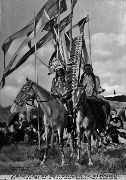 A view of two men on horses with many banners in the background. This was during the adoption of George Miller of the 101 Ranch, one of the early focal points of the oil rush in northeastern Oklahoma, into the Ponca Native American Tribe. Caption reads: "Adoption of Geo. Miller of 101 Ranch into the Ponca Indian Tribe, Oklahoma."