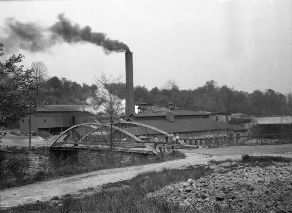 View across road toward a bridge and a factory. There are several boys standing on the bridge.