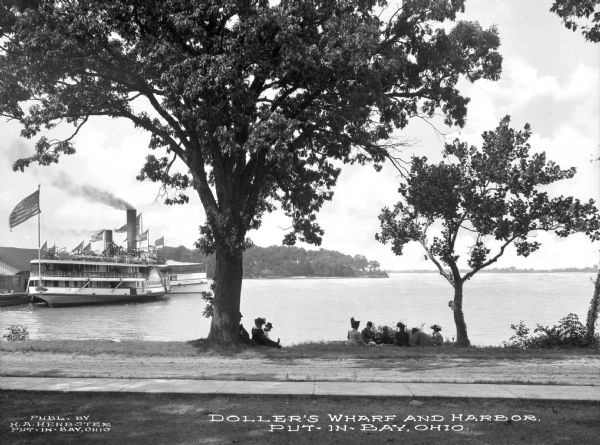 View of a group of men and women sitting on the shore of Doller's Harbor. Nearby, a steamship can be seen at Doller's Wharf. Caption reads: "Doller's Wharf And Harbor, Put-In-Bay, Ohio."