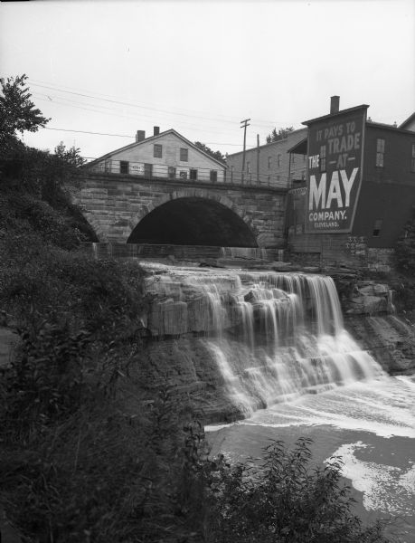 Bridge over the Chagrin river and falls in town with surrounding buildings and an advertisement for the May company.