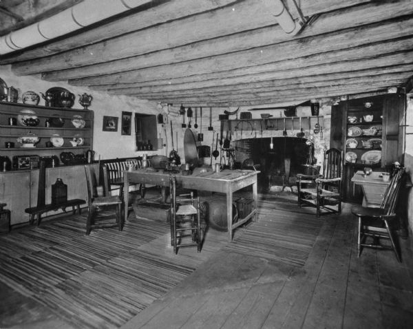 Interior view of the colonial kitchen at the Van Cortlandt house featuring simple wooden table and chairs, a large fireplace for cooking,  cooking implements hanging above the fireplace, and wooden cupboards holding plates, teapots and other ceramic ware.