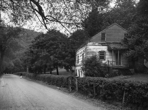 View of an old cabin, built in 1860, on the grounds of the Bedford Springs Hotel. In the foreground is a dirt road, and a pedestrian is walking near the fence.