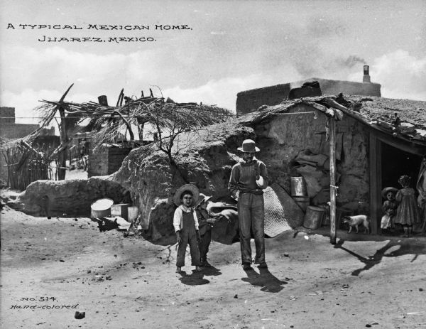 View of man and two boys standing in front of an adobe building. In the background, two girls are playing in the building's entrance while a dog walks by them. The caption reads: "A Typical Mexican Home. Juarez, Mexico."