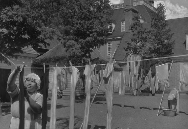 View of two maids in aprons and bonnets hanging laundry out to dry in the yard of the Governor's Palace. The Palace can be seen in the background past trees and outbuildings.