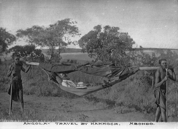 View of a white man reclining in a pole strung hammock, pith helmet by his side. The hammock pole is being carried by two Africans and savannah is in the background. The caption reads "Angola- Travel by Hammock, Mbondo."