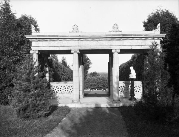 A classical-style stone gazebo with a sculpture on a pedestal at the entrance to a rose garden.