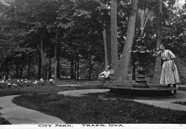 Woman drinking a cup of water from a fountain with a Pan sculpture in City Park. A toddler sits in a nearby baby carriage. The caption reads, "City Park, Traer, Iowa."