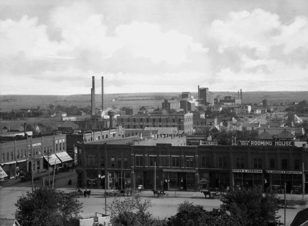 Elevated view of warehouse district east of downtown. Tall brick buildings including the Alton Mercantile Company stand next to wide streets with sidewalks, pedestrians and horse-drawn wagons. The edge of town is in the background.