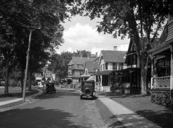 View down tree-lined street, with houses and a sidewalk on the right. Utility lines run the length of the street, and two automobiles are parked at the curb, one on each side of the street.