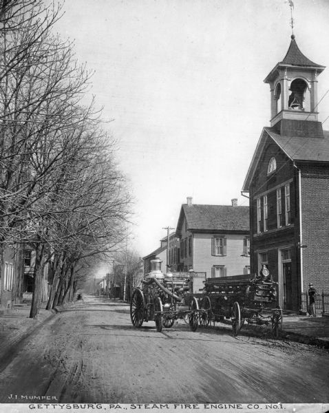 View toward the fire engine which is parked in the middle of the street. To the right, parked next to the curb, is the wagon that carries the fire fighters. Trees line the street on the left side, with buildings and houses on the right. Electric lines run parallel to the street. Caption reads: "Gettysburg, PA., Steam Fire Engine Co. No. 1."