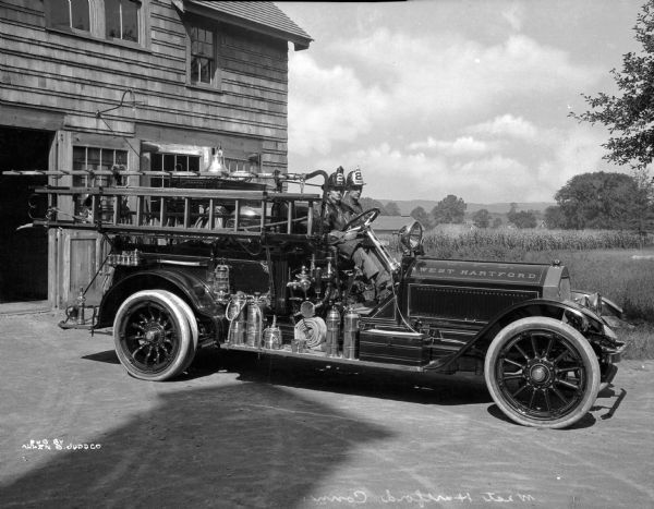 A view of the fire engine, with two firemen posing in the front seat, wearing their uniforms.  The helmets have the number "2" printed on them.  The fire station is visible on the left side, with tress and hills seen in the background, to the right of the scene.  Printed in the bottom left corner: "Pub. by Allen B. Judd Co."