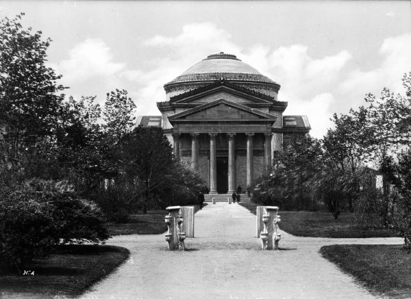 View toward the library's facade, taken from a path leading to the stone and brick building. Two paths intersect the main path and trees and shrubbery line the walkway. The library architecture references the Roman Pantheon and features a Classical-style porch, columns, pediment, and dome.
