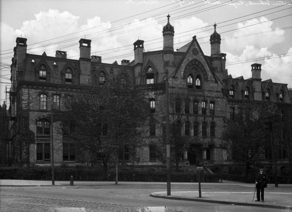 A view of Logan Hall, home to the Wharton School of Business. The building features Gothic Revival elements, including the arched windows and quatrefoils found on the top windows. The main entrance has castle-like details, as shown in the small towers found around the roof line.  A man wearing a top hat and holding a cane stands across the street from the structure and looks at the camera.