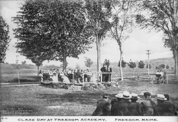 Male and female students from Freedom Academy sit outdoors with two teachers. A larger group sits on platform while a smaller group looks on from the foreground. Caption reads: "G-11. Class Day at Freedom Academy. Freedom, Maine."