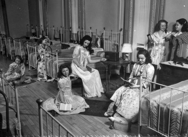 Female students wearing pajamas and robes sit reading or talking with one another in what appears to be a dormitory room at Holy Family Academy. Beds line the walls and a chest of drawers or a table stands next to each one.