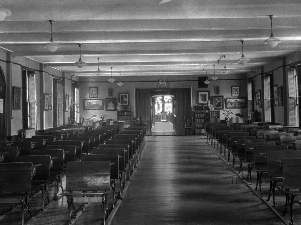 View down aisle of the study hall at St. Joseph's Academy of Maine. Bookcases line the back walls and are decorated with bust sculptures. Framed artwork hangs on the walls and a crucifix stands in the background.