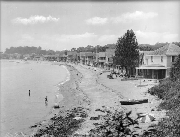 Elevated view of a beach and houses that line the shore. People sit on the beach, and a woman in the foreground wades in the water.