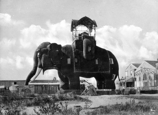 A view of the left side of the Elephant Hotel, constructed in the shape of an elephant with small windows punctuating the side and a covered open-air observatory at the top. The hotel stands in a sandy area and various buildings are behind it. The hotel is also known as Lucy the Elephant and was built by James V. Lafferty in 1882.