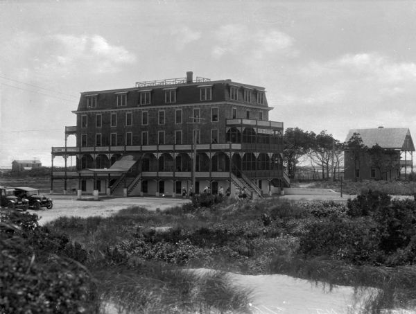 View across sandy beach with grasses toward a group of people standing near the staircase to what appears to be a large boarding house or hotel. The first and second stories have porches that run their length and a sign reading "Sun-Set" is affixed to the right side of the building.  Multiple automobiles are parked in a dirt lot to the left.