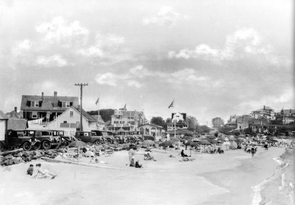 View along beach toward sunbathers sitting and walking along the seashore. A line of automobiles are parked alongside the beach, separated from the sand by a low rock wall. In the background are businesses and summer homes.