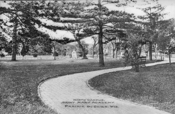 View across curving brick walkway in the lawn of North Campus at St. Mary Academy. A few structures are in the background, and a bench stands near the path. Caption reads: "North Campus, Saint Mary Academy Prairie du Chien, Wis."