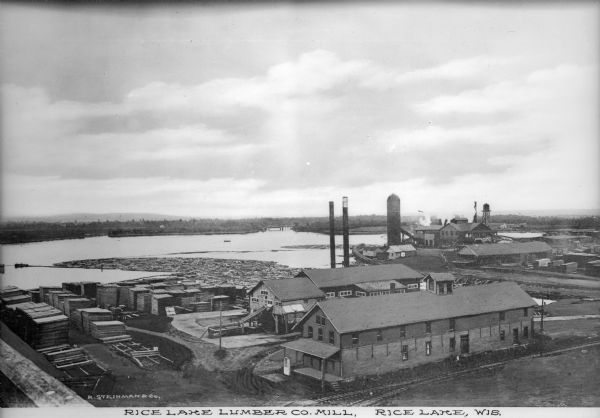 Elevated view of the Rice Lake Lumber Company Mill, taken from a high vantage point. A formation of logs is in the river, and piles of finished boards are in the foreground. Caption reads: "Rice Lake Lumber Co. Mill, Rice Lake, Wis."