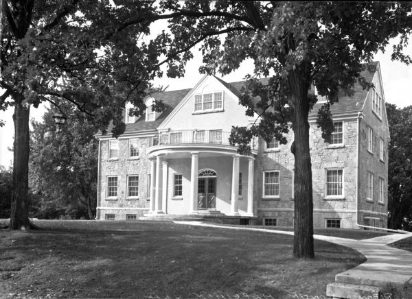 A pathway leads to Merriman Hall at Ripon College. The columned porch shelters a double door entrance and supports a second floor balcony.  The middle third of the structure appears to be covered in plaster, while the sides are stone.