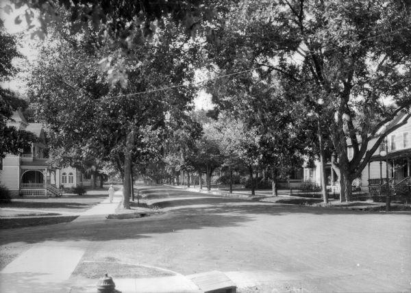 View across intersection toward houses on both sides of a tree-lined street. A woman is walking down the sidewalk on the left side of the street, and another woman is sitting on the porch of a residence on the right side of the street.