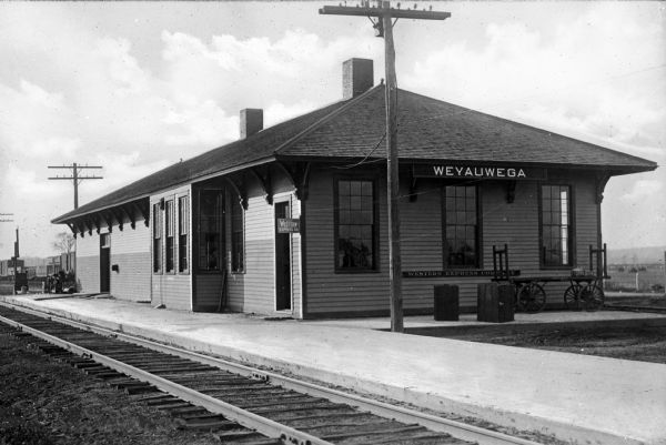 A view of the Weyauwega Train Depot building, platform, and railroad tracks. Signs on the building read, "Agency Western Express Co." and "Western Express Company."