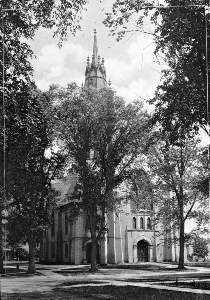 A view of the facade and side of the First Methodist Church. All visible windows are made of stained glass and the highly ornate steeple is partially obscured by trees.