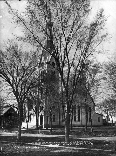 View of the  facade and side of the Congregational Church, partially obscured by trees. The bell tower doubles as a clock tower.