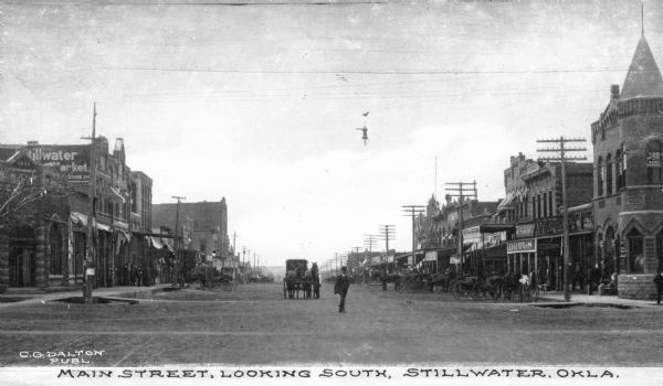 A view of a broad street lined with businesses in brick buildings. Horse-drawn carts are parked along the street and pedestrians are visible as well. Signs read: "Pathfinder Cigars" "Coca-Cola" and "Law Office of Bush & Bowers." Caption reads: "Main Street, Looking South, Stillwater, Okla."