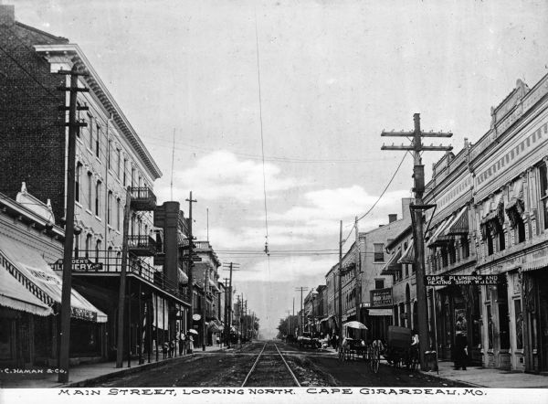 Electric cable car tracks run down the street and horse-drawn carts are on the right side of the street. Electrical power lines are strung across the street. Business signs read: "Cape Plumbing And Heating Shop. W.J. Lee" and "Restaurant Arcade Saloon Oyster Cafe." Caption reads: "Main Street, Looking North, Cape Girardeau, Mo."