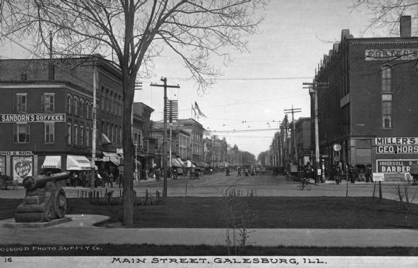 View of the Main Street shopping district. Storefronts, carts and pedestrians are across a small grassy area with a military monument of a cannon. Signs read: "Sanborn's Coffees" "Gordon & Bennetts A Royal Slave" "Raymond & Sons" "Schloss Clothing" "Socialist Club" "Foster's" "Miller's" and "Ingersoll." Caption reads: "Main Street, Galesburg, Ill."
