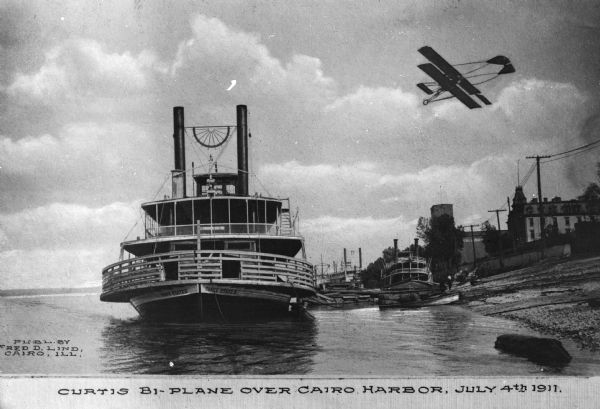 View of a Curtiss biplane flying over the harbor. The steamship "Three States" are in the center, and several similar ships are in the distance, including one bearing the letter "B." Caption reads: "Curtis Bi-Plane Over Cairo Harbor, July 4th, 1911."
