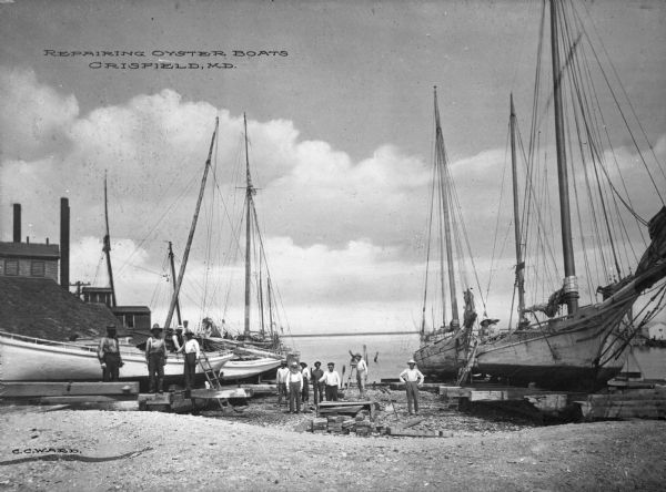 View of a group of people posed near several oyster boats under repair on the shore. Some buildings are on the left side. A sign on the boat on the far right reads: "Russell." Caption reads: "Repairing Oyster Boats Crisfield, M.D."