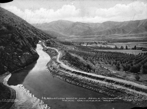 View of an irrigation ditch near apple orchards in. Mountains are in the distance. Caption reads: Irrigation Ditch And Apple Orchards, Near Cashmere, Wash."
