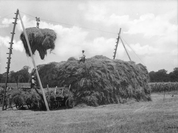 Two agricultural workers use machinery to stack alfalfa in the vicinity of Tulsa. One man stands on the large pile while another uses a pulley device to stack the alfalfa. A cornfield is in the background.