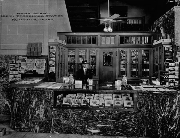 A man working behind the counter of a very elaborate and ornate newsstand in a Union Railroad Passenger Station. Caption reads: "New Stand, Union Passenger Station, Houston, Texas."