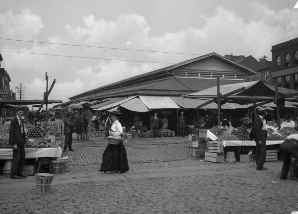An outdoor open-air Lexington market in Baltimore, MD. Fruit and produce stalls can be seen on on either side of the picture and in the background a large covered structure is also visible. A woman in a long skirt and hat is carrying a basket in the center of the frame.