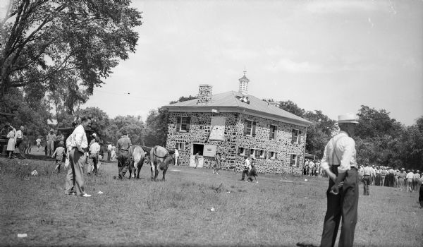 A crowd has gathered at a well preserved large stone barn with a cupola.  Two men with cameras watch the scene from a hatch in the roof; there is a van with "Chicago (?) Tribune" painted on the side, and a sign on the barn door reads "Press."  A man drives a pair of yoked oxen toward the barn.