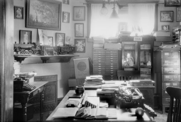 A view of a cluttered office, possibly that of the photographer, with several desks and two typewriters. There is a large model train on a mantle above a small fireplace. There are prints of female nudes on the walls.
